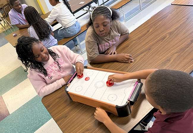 Two students play a air hockey game as a girl looks on.